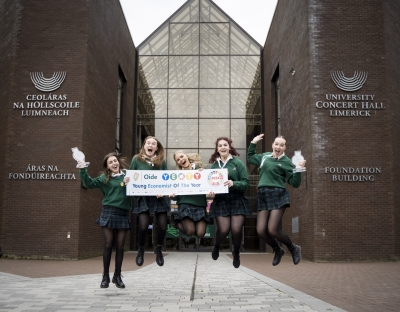 Five very happy teenage girls in green secondary school uniforms jumping for joy holding a poster which reads Young Economist of the Year